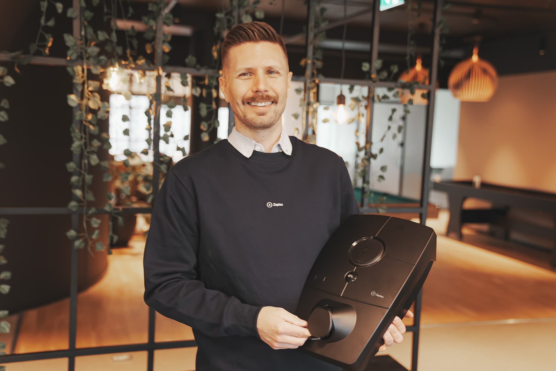 En smilende mann i en marineblå genser holder en Zaptec Pro-lader i et moderne kontorlokale med varm belysning, hengende grønne planter og treelementer.