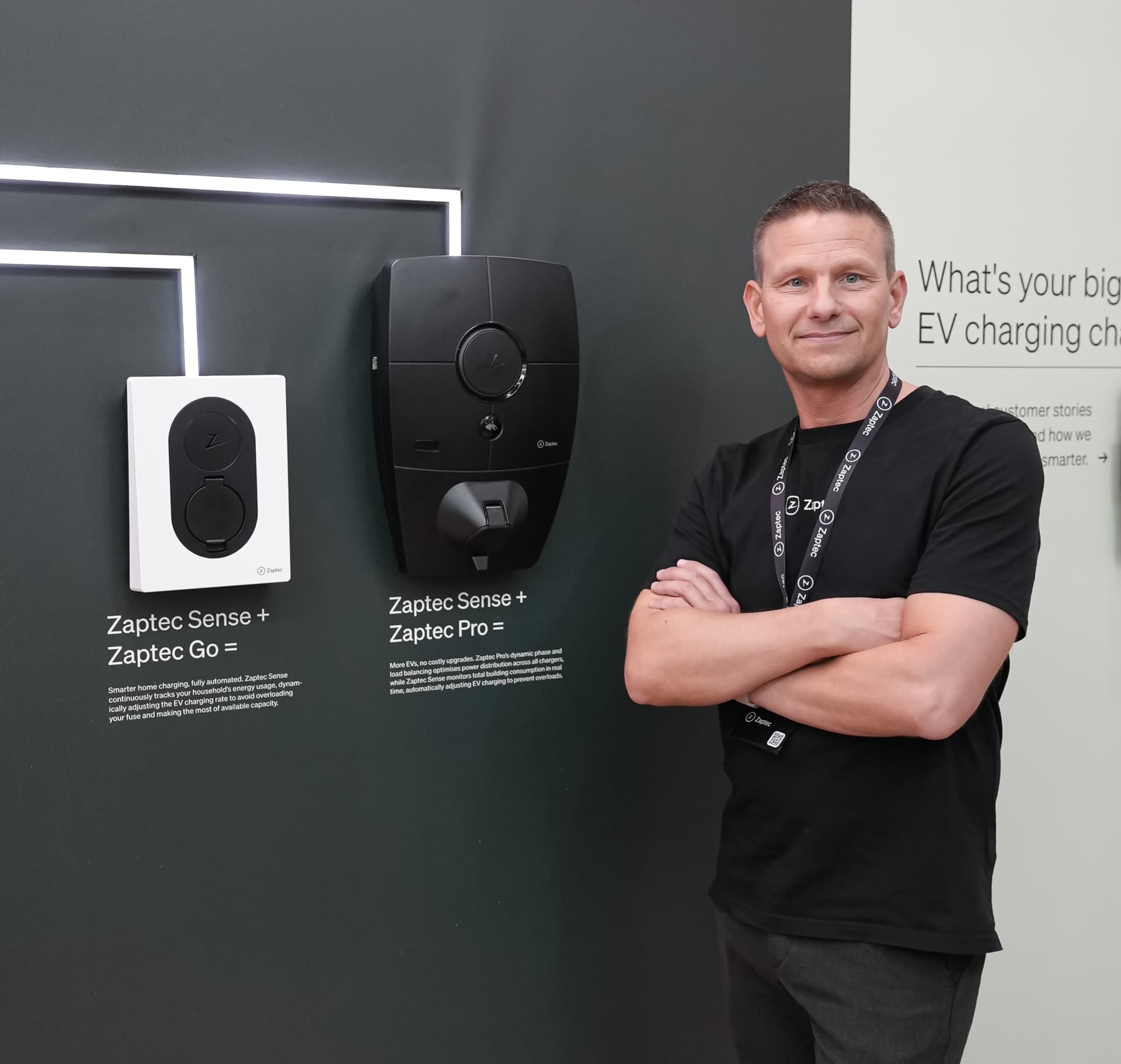 A smiling man stands next to a display wall showing a Cloud White Zaptec Go and an Zaptec Pro. The display highlights the different models and their design features.
