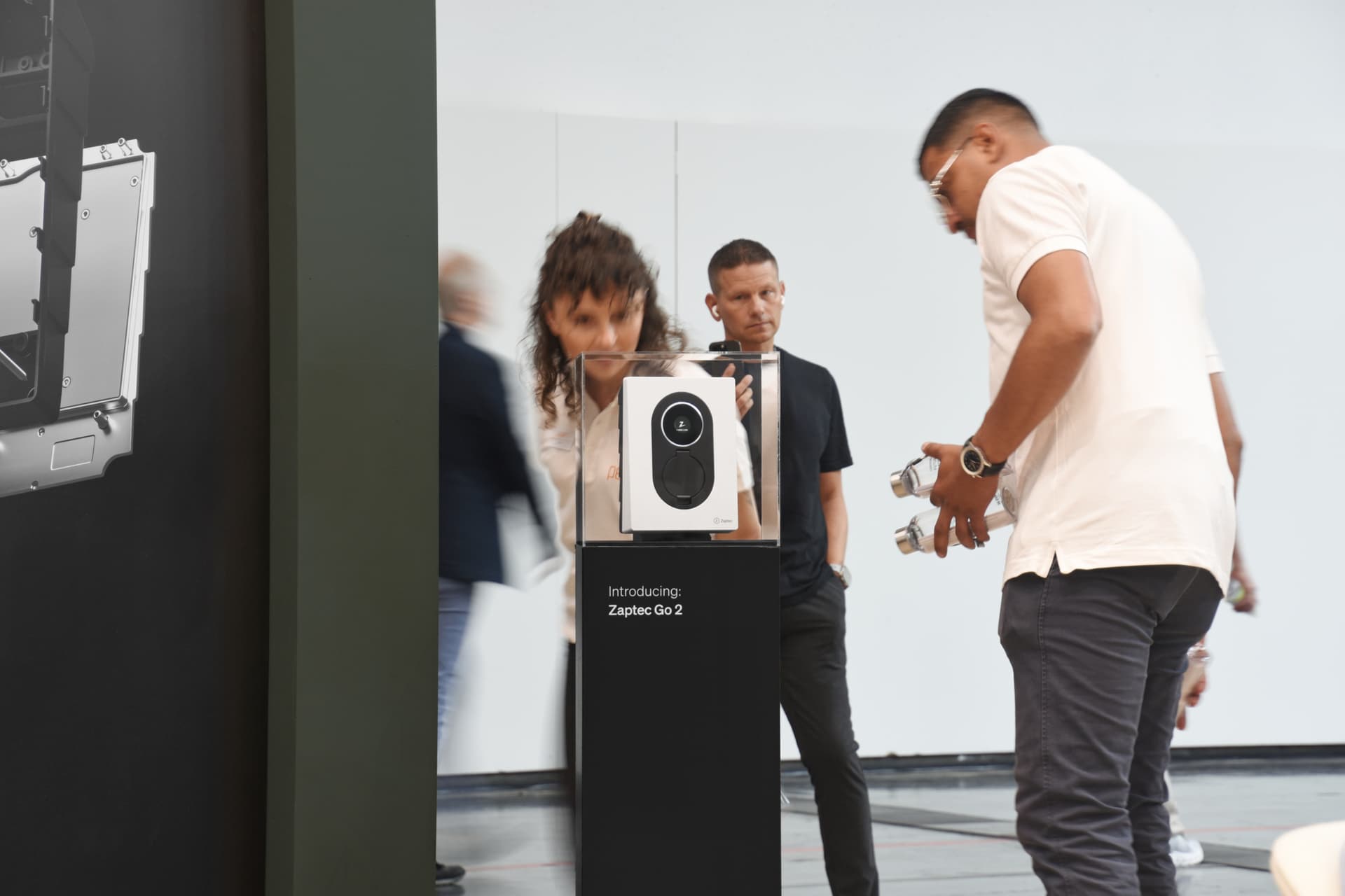 En Cloud White Zaptec Go 2-lader står utstilt på en svart søyle i et lyst og moderne innemiljø. Flere personer studerer laderen på nært hold.