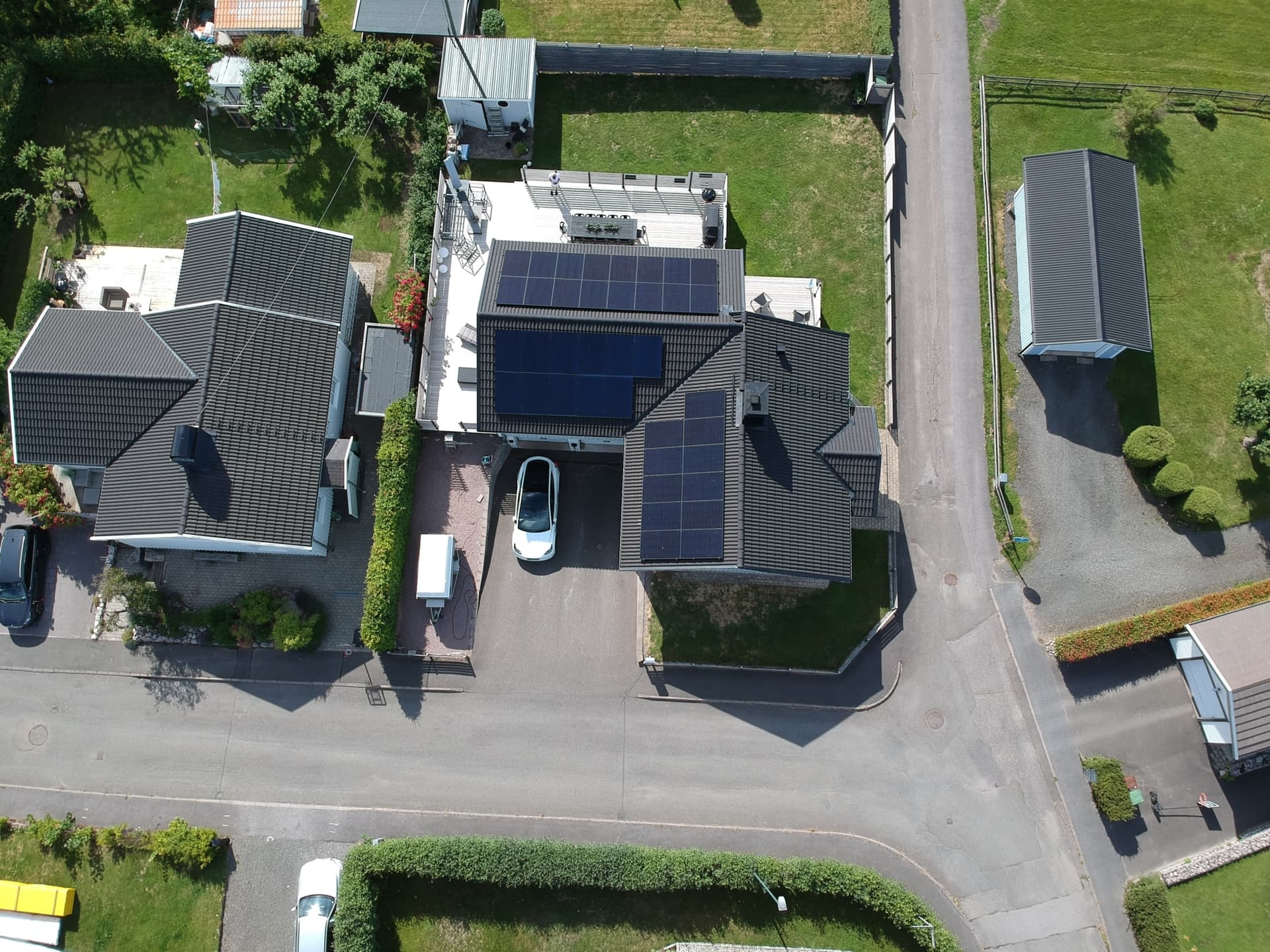 Aerial view of a white house with solar panels on the roof and a white electric car parked in the driveway.