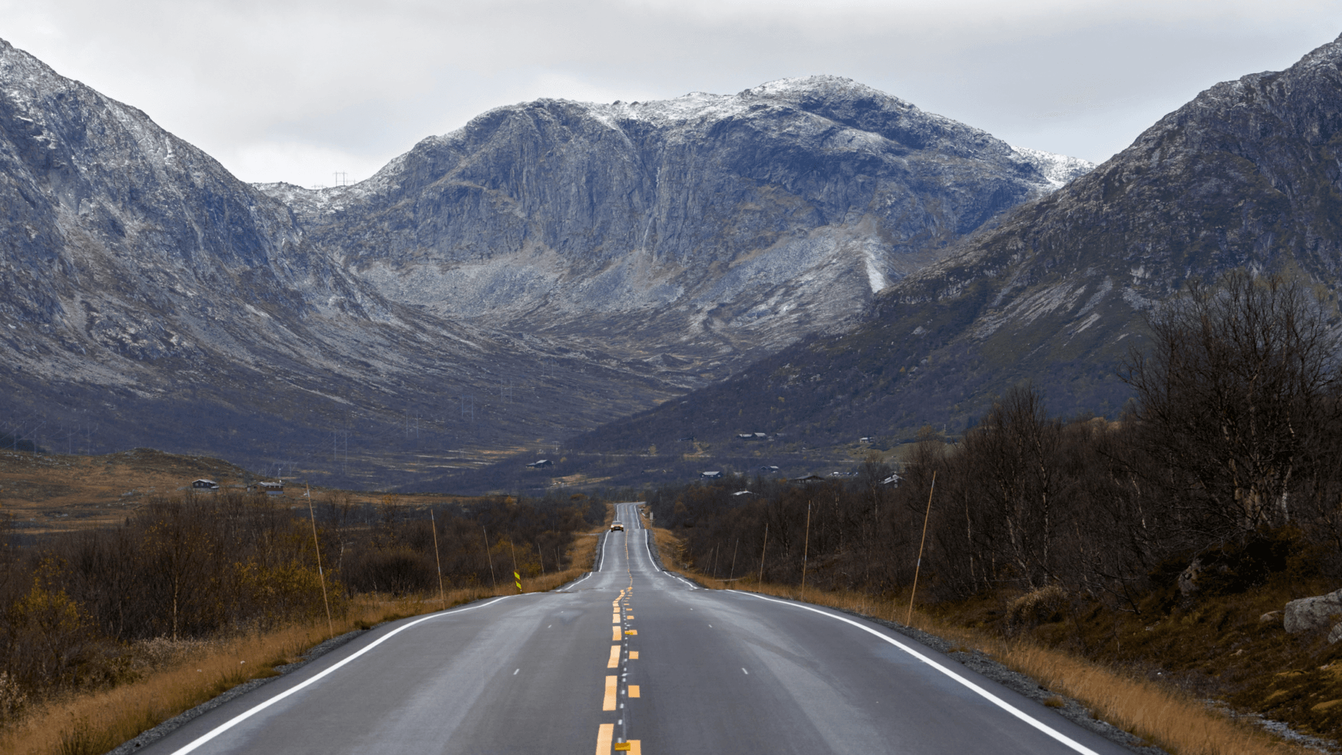 A long, empty road stretches toward dramatic mountains with snow-covered peaks under a cloudy sky. Autumn colors line the road, creating a calm feeling before winter.