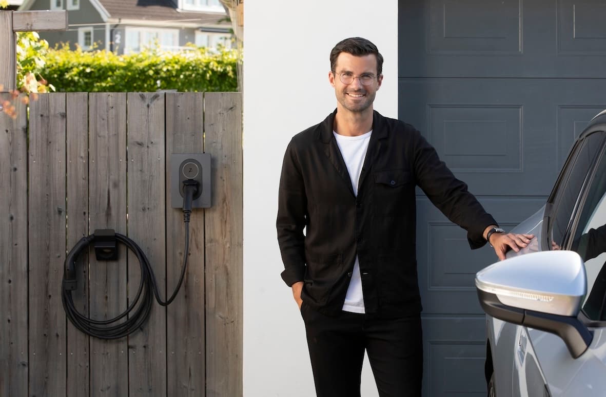 John Wastring Zaptec Sverige, står med svart knäppt overshirt, vit t-shirt, glasögon och mörka byxor vid en silvergrå elbil, i bakgrunden är ett modern garage i vitt med grå port. På ett träplank i grå nyans bredvid garaget sitter en går Zaptec Go med en laddkabel prydligt ihoprullad och upphängd på kabelhållare Zaptec Chill