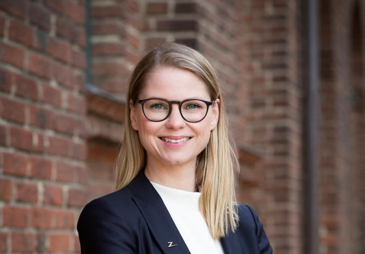 En kvinna med blont hår, glasögon och en marinblå kavaj står leende framför en tegelvägg