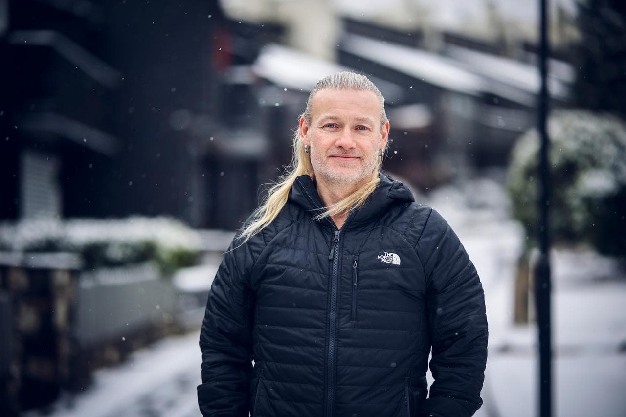 En mann med langt blondt hår og svart jakke står utendørs i et boligområde dekket av snø, med snøfnugg som faller rundt ham.