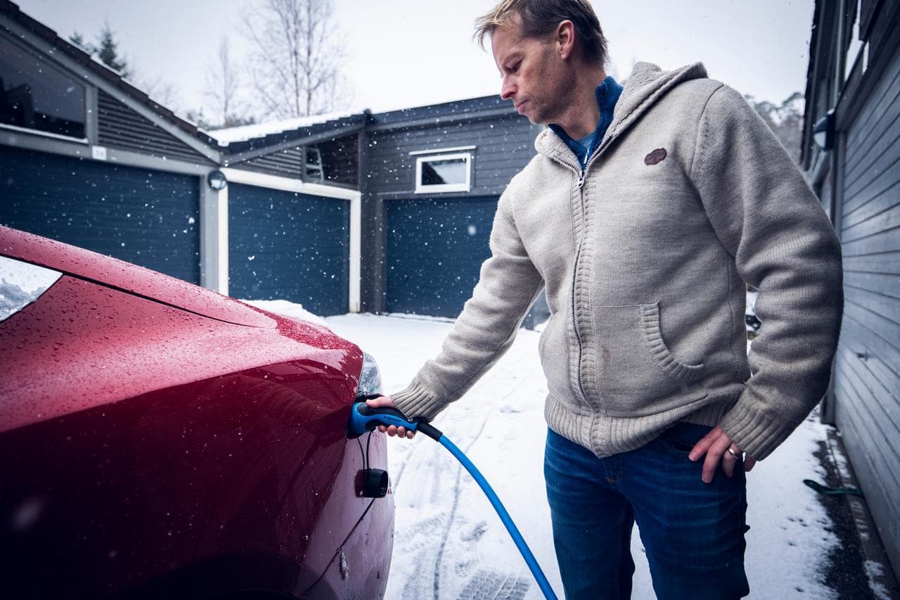 En man i beige tröja och jeans kopplar in en blå laddkabel i en röd elbil. Runt omkring syns ett snöigt landskap och garageportar i blå grå nyans.