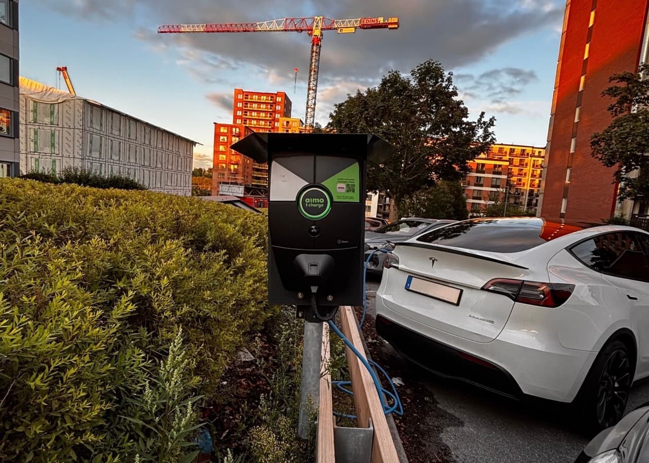En vit elbil står parkerad och laddas med en Zaptec Pro-laddare monterad på en stolpe i en bostansmiljö. Laddaren är delvis skuggad av ett tak, och i bakgrunden syns bostadshus och grönska i solnedgången.