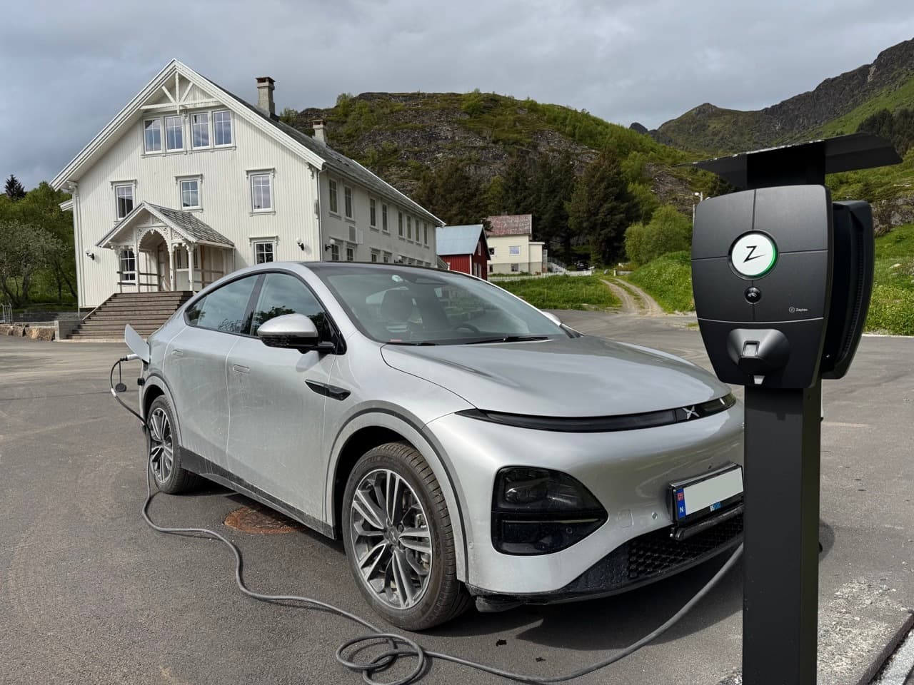 En sølvfarget Xpeng G9 elbil står til lading ved en Zaptec Pro lader som er montert på stolpe, foran et stort hvitt trehus omgitt av grønn natur og fjell.