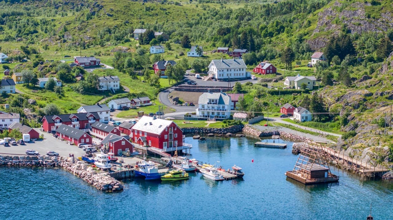 Fiskerihavn med småbåter ved brygge, røde naust og hus, omgitt av grønn vegetasjon og fjell i bakgrunnen