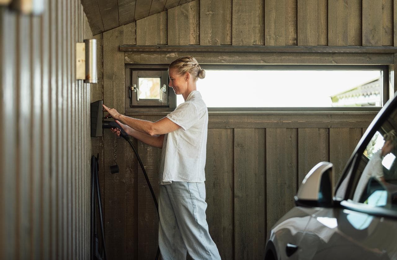 En kvinna med ljusa luftiga kläder sätter i en laddkabel i en Zaptec Go laddbox i ett garage med väggar i grått trä.