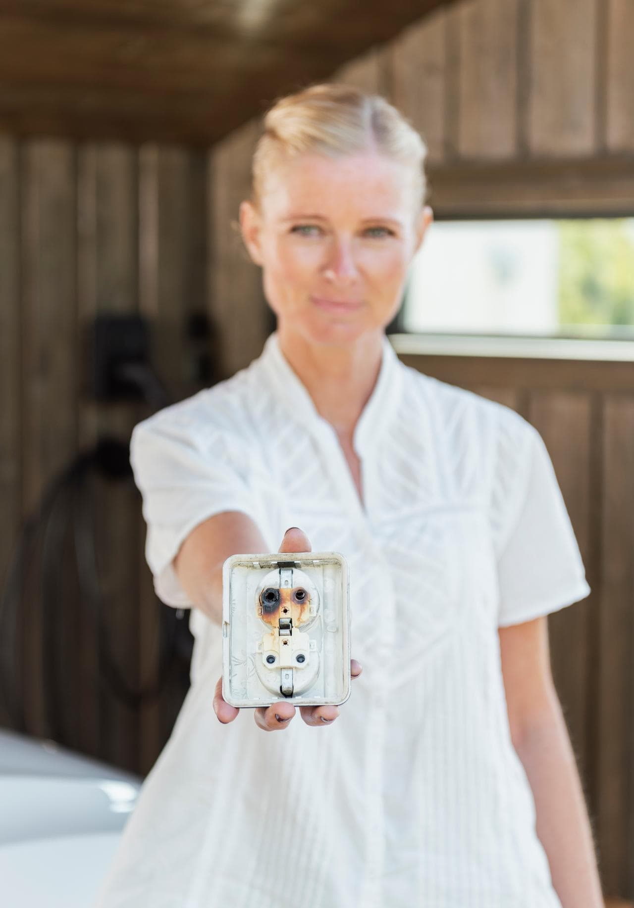 En kvinna med ljusa luftiga kläder och blont uppsatt hår håller fram ett eluttag som är nermonterat från väggen, det syns tydligt att det har blivit bränt i uttaget, kvinnan står ett garage med väggar i grått trä.
