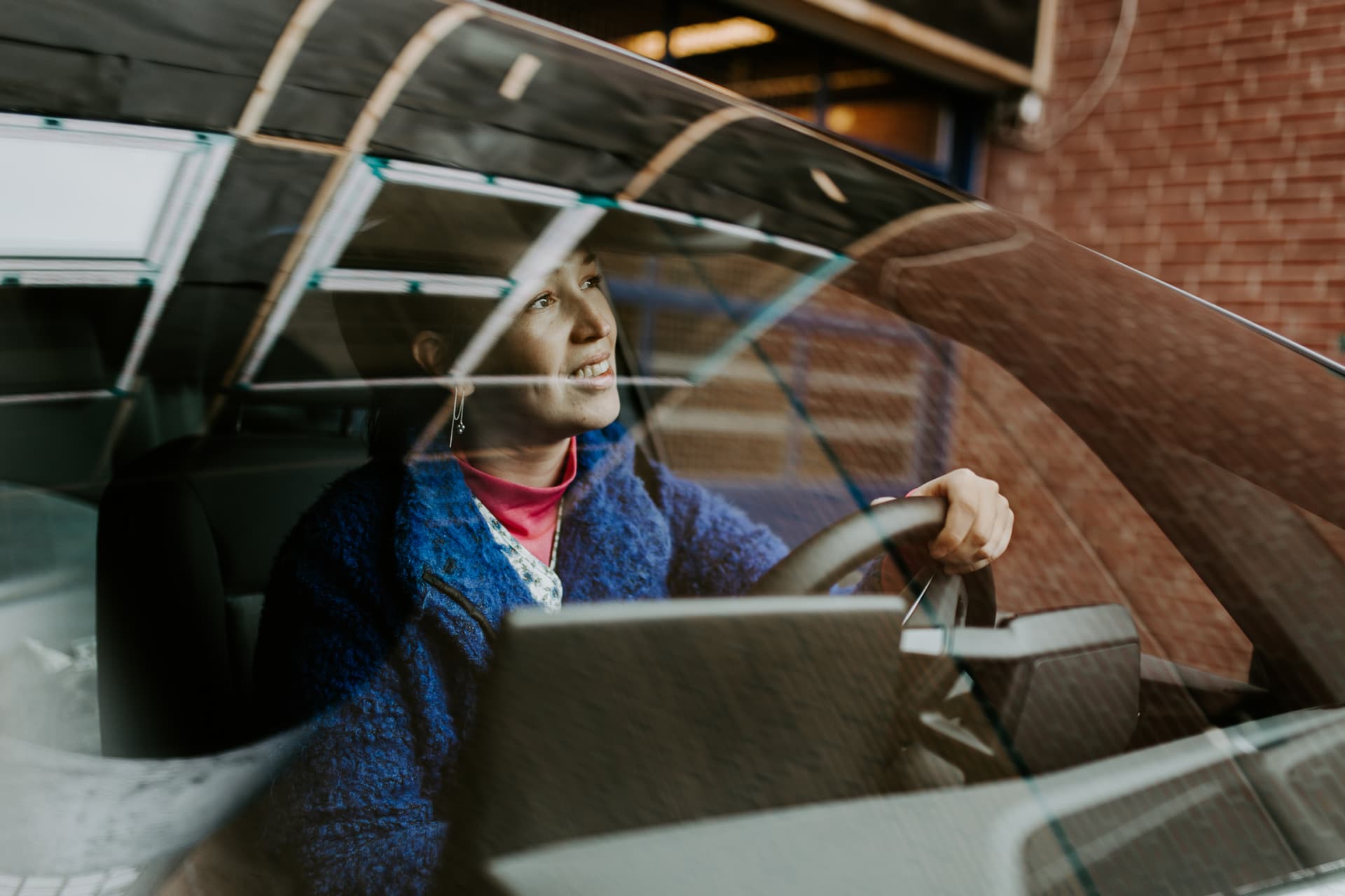 genom framrutan på en bil syns en kvinna som leende kollar ut med en hand på ratten, hon är klädd i en blå kappa och röd tröja.