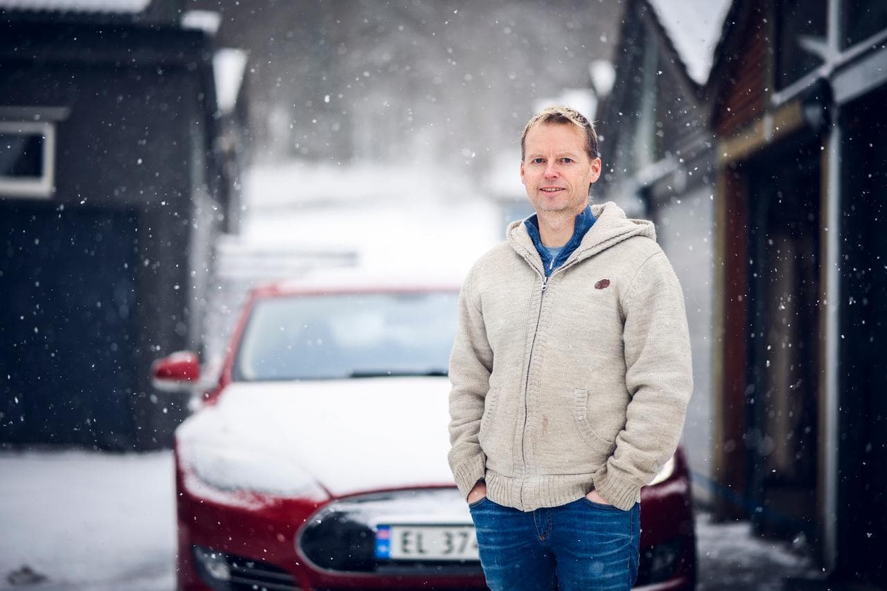 En mann i lys hettegenser står i snøvær foran en rød elbil parkert mellom hus i et vinterlandskap.