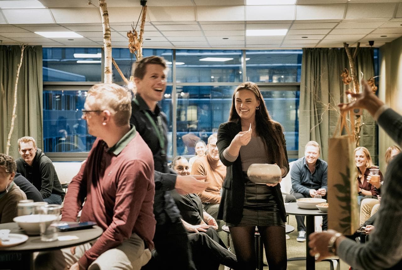 En glad gruppe kolleger er samlet innendørs på et Zaptec-arrangement. En kvinne trekker en lapp fra en bolle mens de andre ler og ser på – det er en livlig og varm stemning.