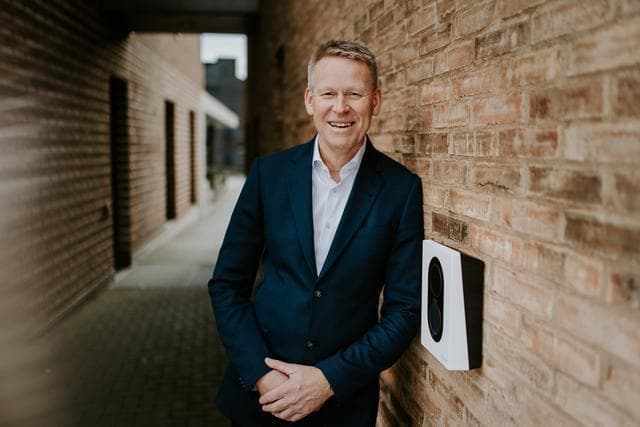 En smilende mann i en mørkeblå dress lener seg mot en murvegg med en Cloud White Zaptec Go-lader montert på den.