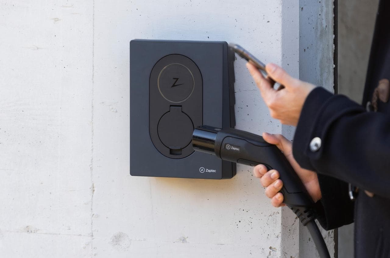 En person holder en ladekabel og en mobiltelefon foran en Asphalt Black Zaptec Go-lader som er montert på en hvit vegg.