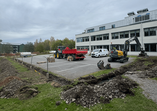 Förberedelse för installation av laddboxar på en parkeringsplats med grävmaskin, traktor och en vit skåpbil står parkerad vid kontorsbyggnaden.