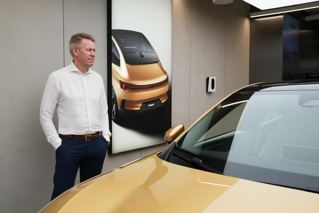 En mann i hvit skjorte står ved siden av en gullfarget elbil i et showroom. På veggen bak ham er en Cloud White Zaptec Go-lader montert ved siden av en plakat med en bil i samme farge.