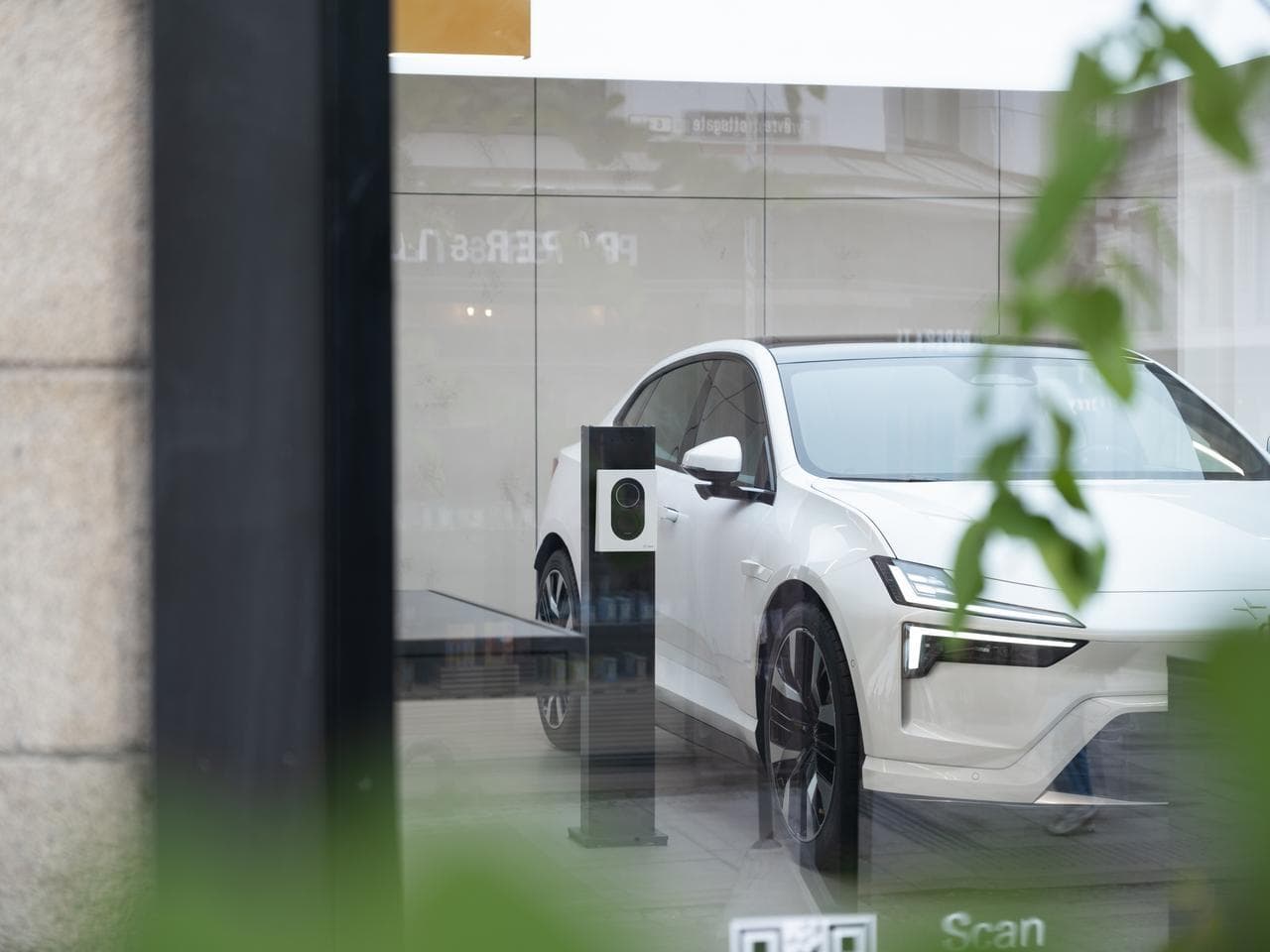 En hvit elbil står parkert innendørs og lader med en Cloud White Zaptec Go-lader, sett gjennom et glassvindu med grønne planter i forgrunnen.