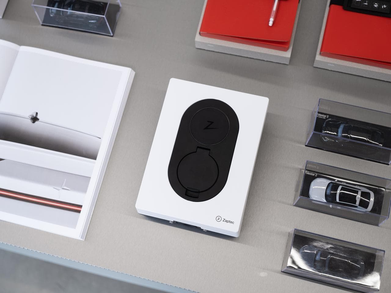 Sur une table grise repose une Zaptec Go blanche entourée de miniatures de voitures ainsi que d'une brochure montrant une voiture électrique blanche.
