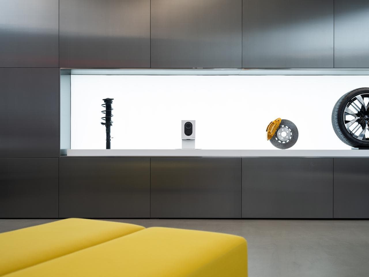 En Cloud White Zaptec Go-lader er utstilt i en opplyst monter sammen med bildeler i et moderne showroom med mørke veggpaneler og en gul sittemøbel i forgrunnen.