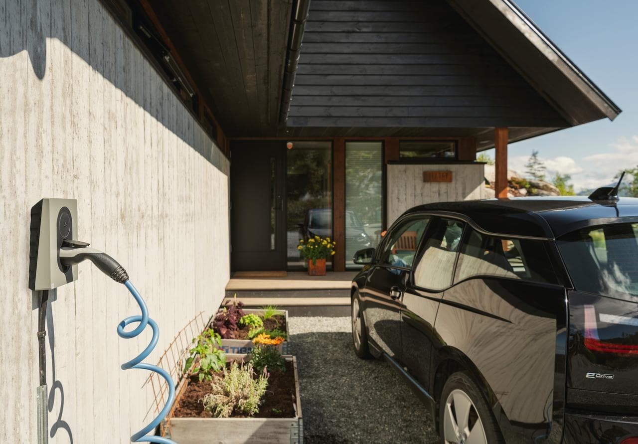 En svart elbil lader med en Moss Green Zaptec Go-lader som er montert på en betongvegg ved et moderne hus med trefasade og plantekasser foran.