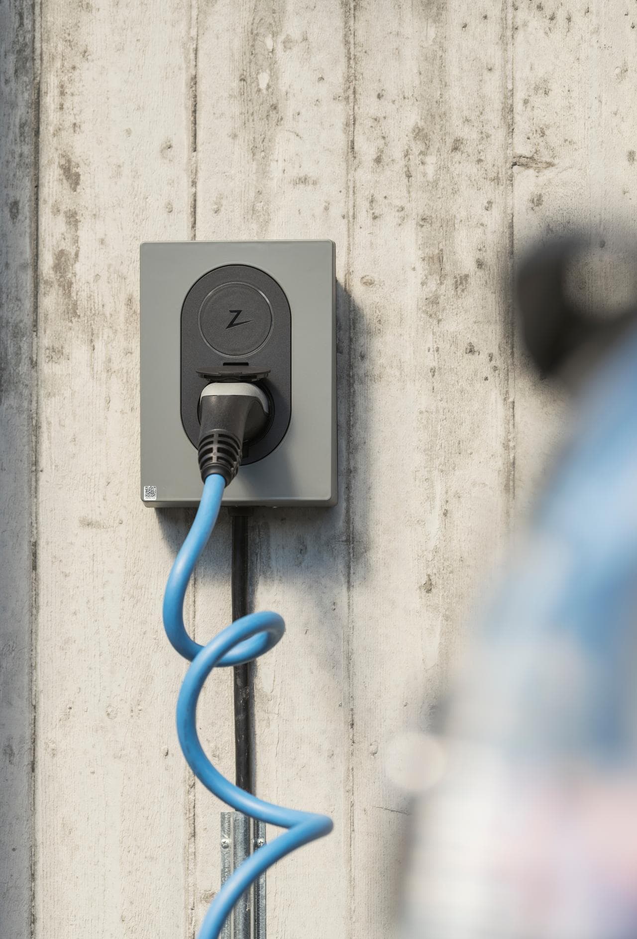 en Moss green Zaptec Go sitter på en betongvägg, med en blå laddkabel inkopplad i elbilsladdaren, längs högra sidan av bilden är sidan av en svart elbil som är ur fokus