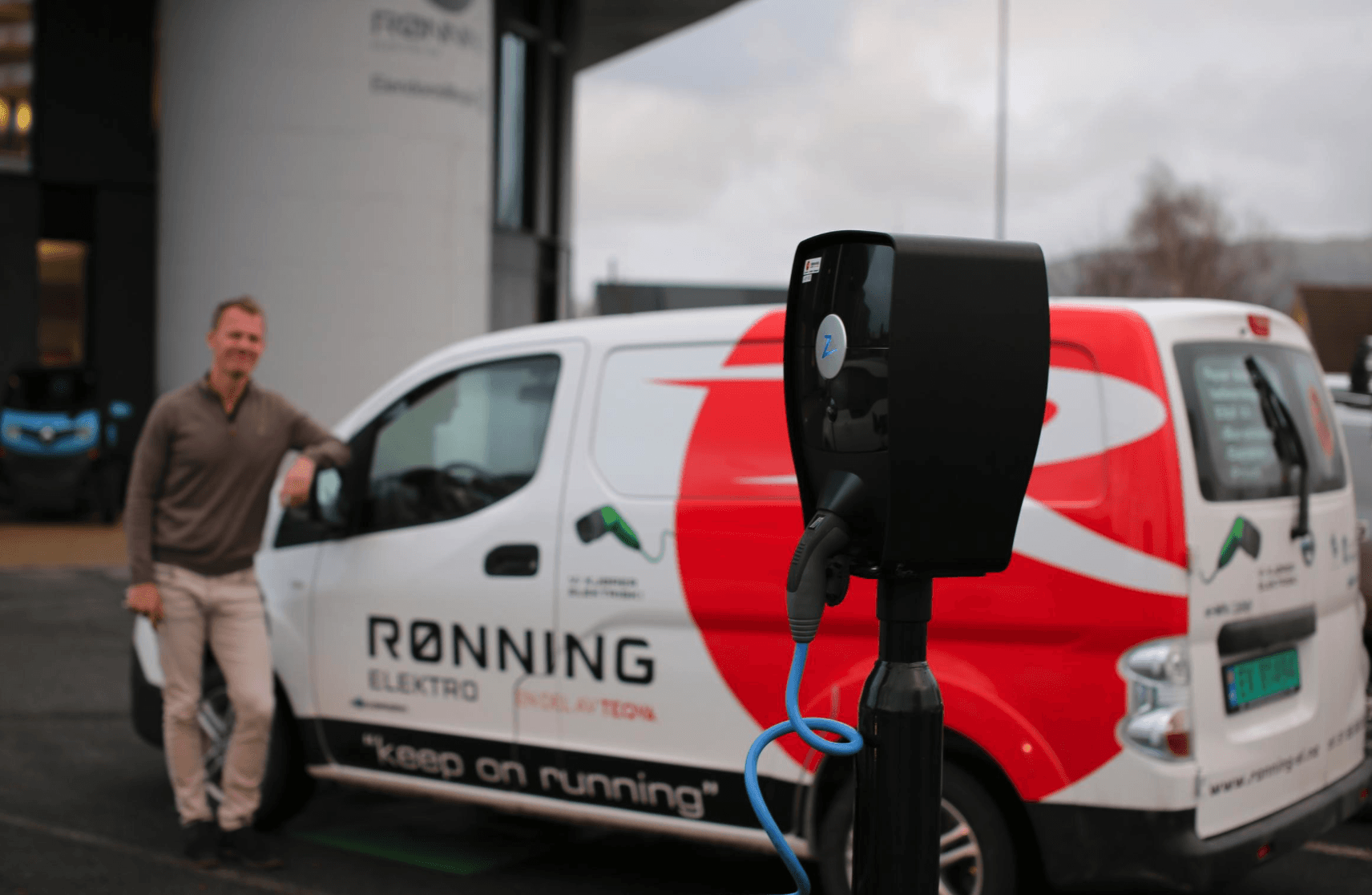 En man står bredvid en vit och röd skåpbil med texten "Rønning Elektro" på, medan en laddningsstation för elbilar från Zaptec syns i förgrunden.