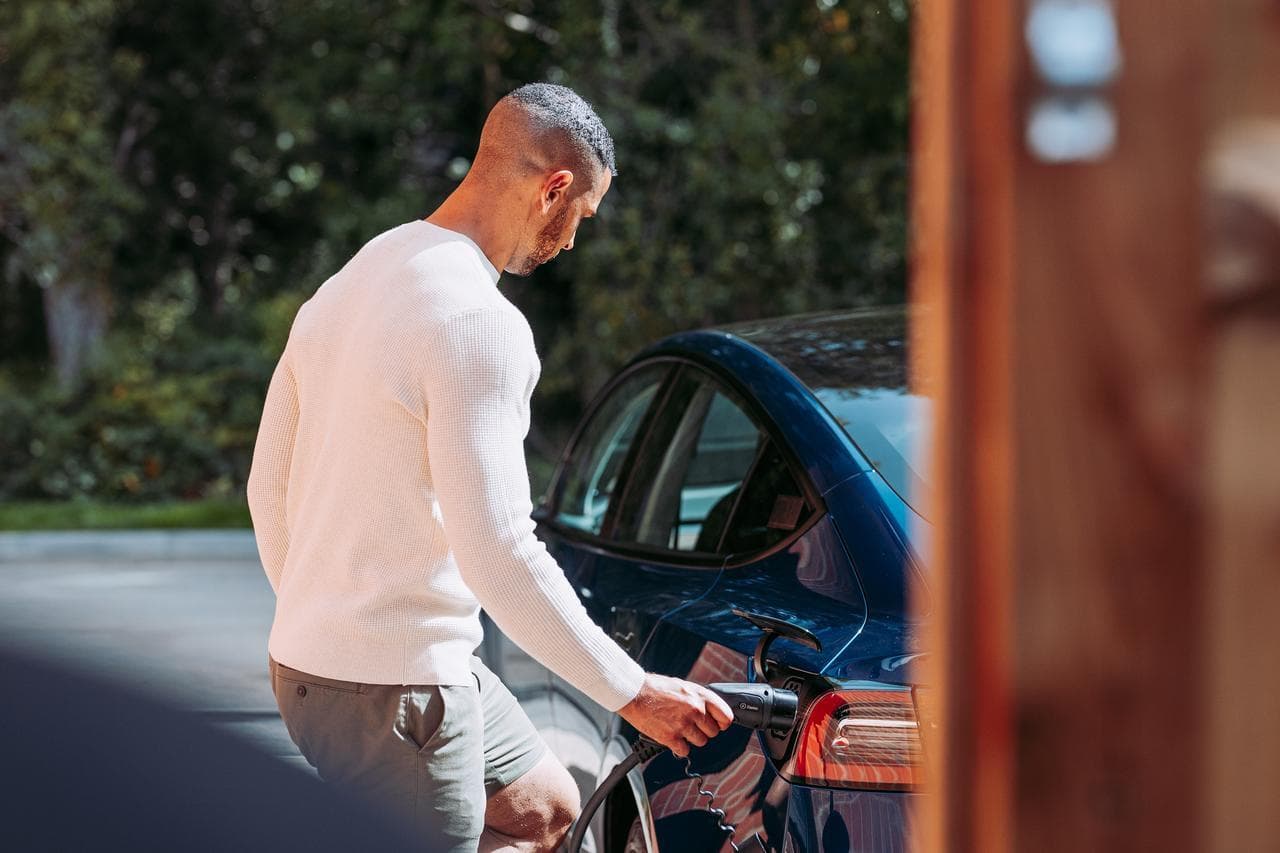 A man in a white sweater connects a charging cable to a blue electric car in a sunny outdoor setting with trees in the background.