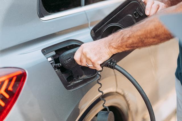 En hånd som kobler til en ladekabel i en sølvfarget elbil, mens sollyset reflekteres mykt på bilens overflate.