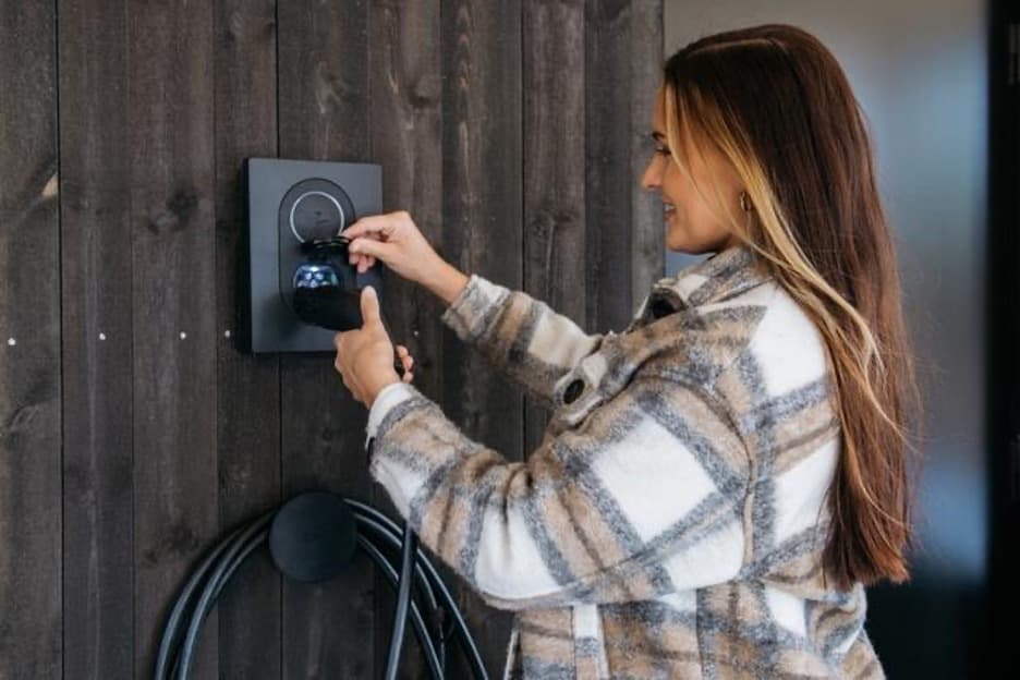 En smilende kvinne i en rutete jakke kobler til en ladekabel i en Rock Grey Zaptec Go-lader som er montert på en mørk trevegg.
