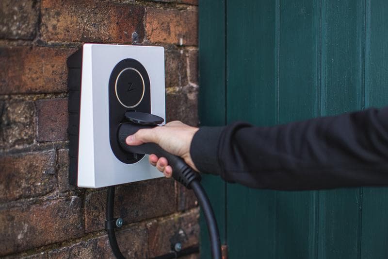 En person sätter i en laddkabel i en vit Zaptec Go laddbox som sitter monterad på en tegelvägg.