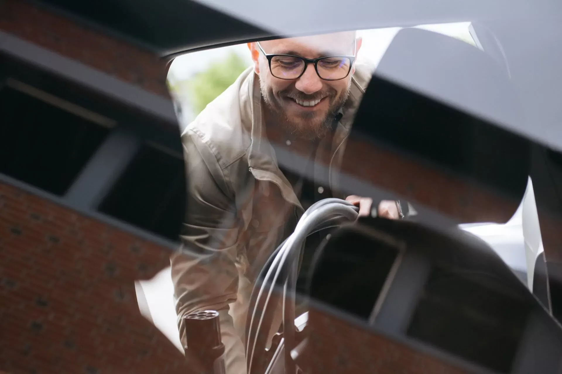 En mann med briller og beige jakke smiler mens han holder i en ladekabel, sett gjennom bilens sidespeil og karosseri.