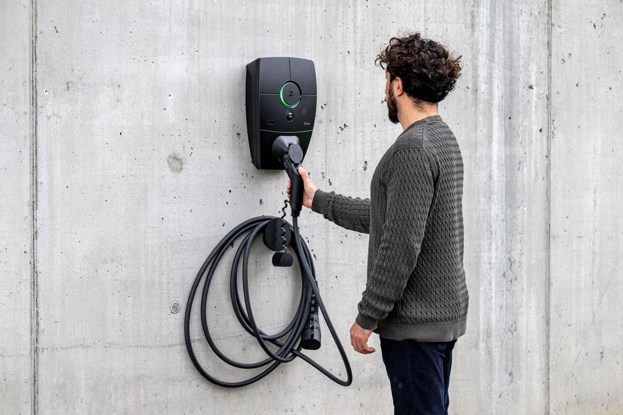 En mann i grå strikkegenser holder en ladekabel koblet til en Zaptec Pro-lader montert på en grå betongvegg.