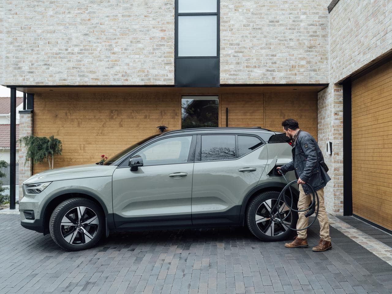 En man med snart jacka och beiga byxor kopplar in en laddkabel från Zaptec i sin grön grå elbil i suv modell, bilen står framför ett modern hur i beige med grå långsmala plattor på marken.