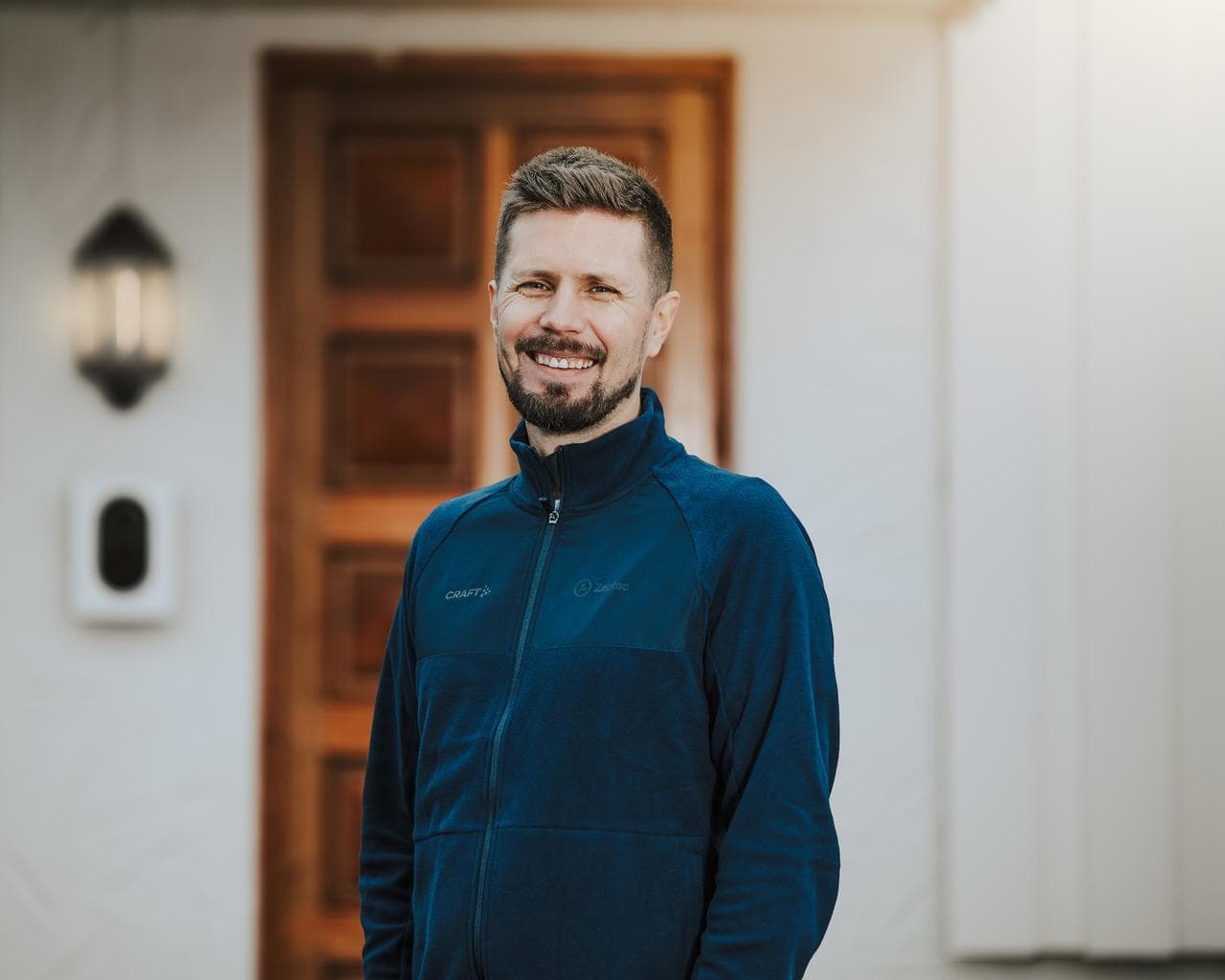 En leede man mad mörkt hår, skägg och blå tröja står framför en vit byggnad med en Zaptec go monterad på väggen.
