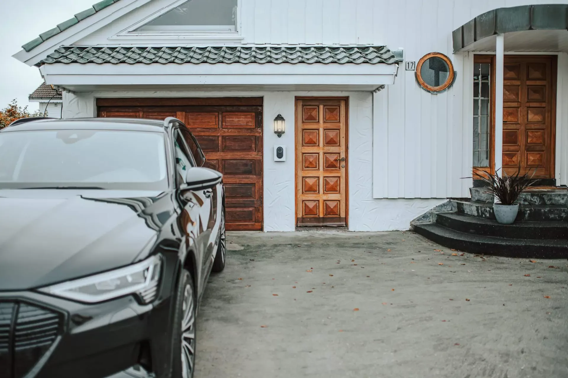 En svart elbil står parkert foran et hvitt hus med tredetaljer og en Cloud White Zaptec Go-lader montert mellom garasjeporten og ytterdøren.