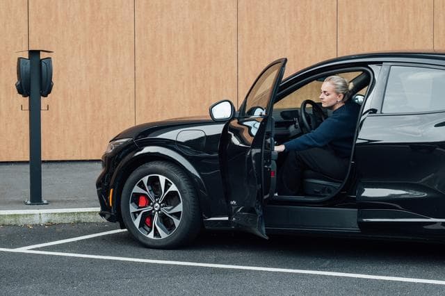 En kvinne sitter i førersetet på en svart elbil med åpen dør, parkert ved siden av en Zaptec-ladestasjon montert på en svart stolpe. I bakgrunnen er en vegg med trepaneler.