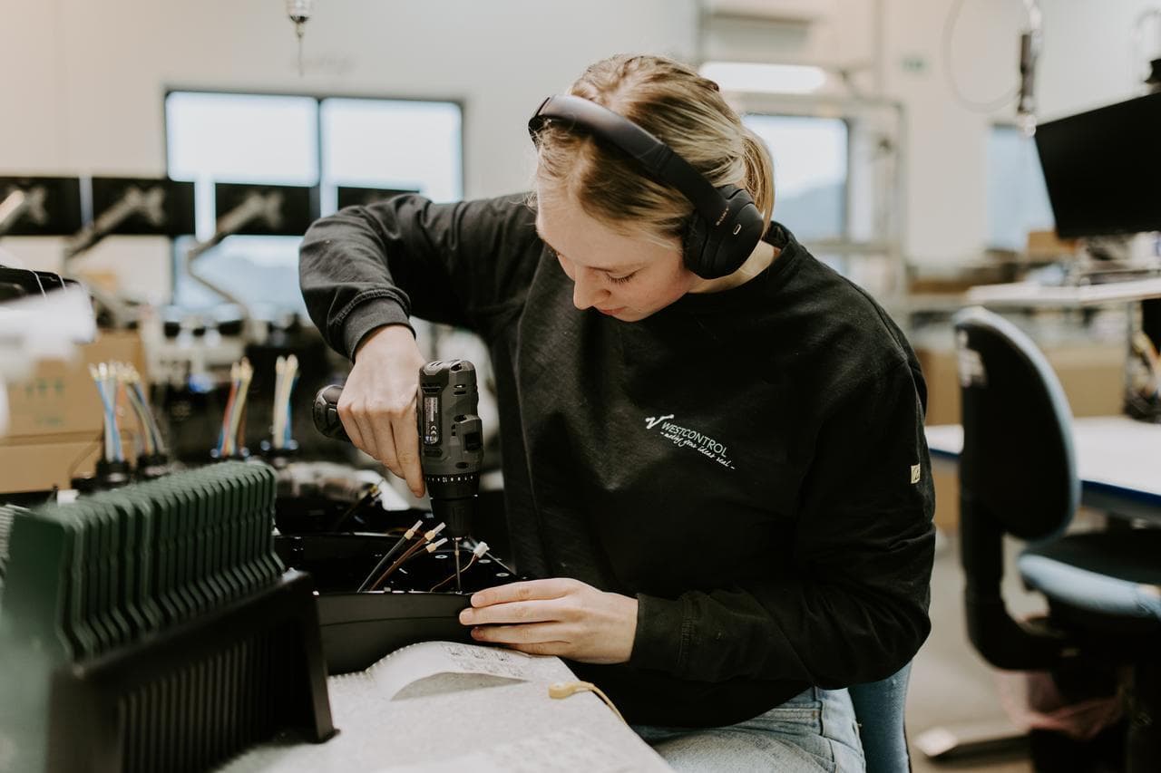 En person med hørselsvern og svart genser jobber konsentrert med montering av elektronikk i et fabrikksmiljø.