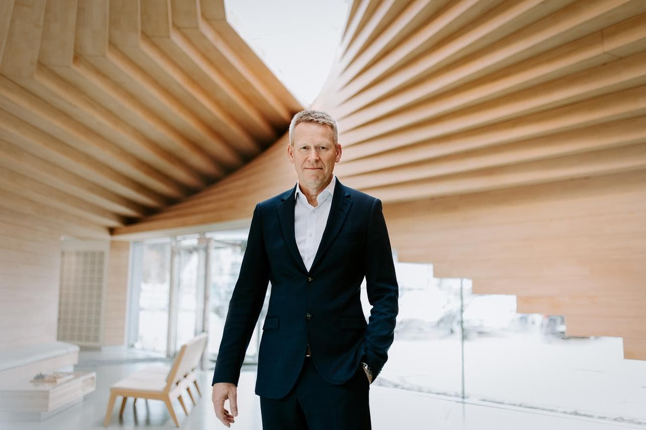 A man in a dark suit standing confidently inside a modern wooden building with dramatic architectural lines and large windows letting in winter light.