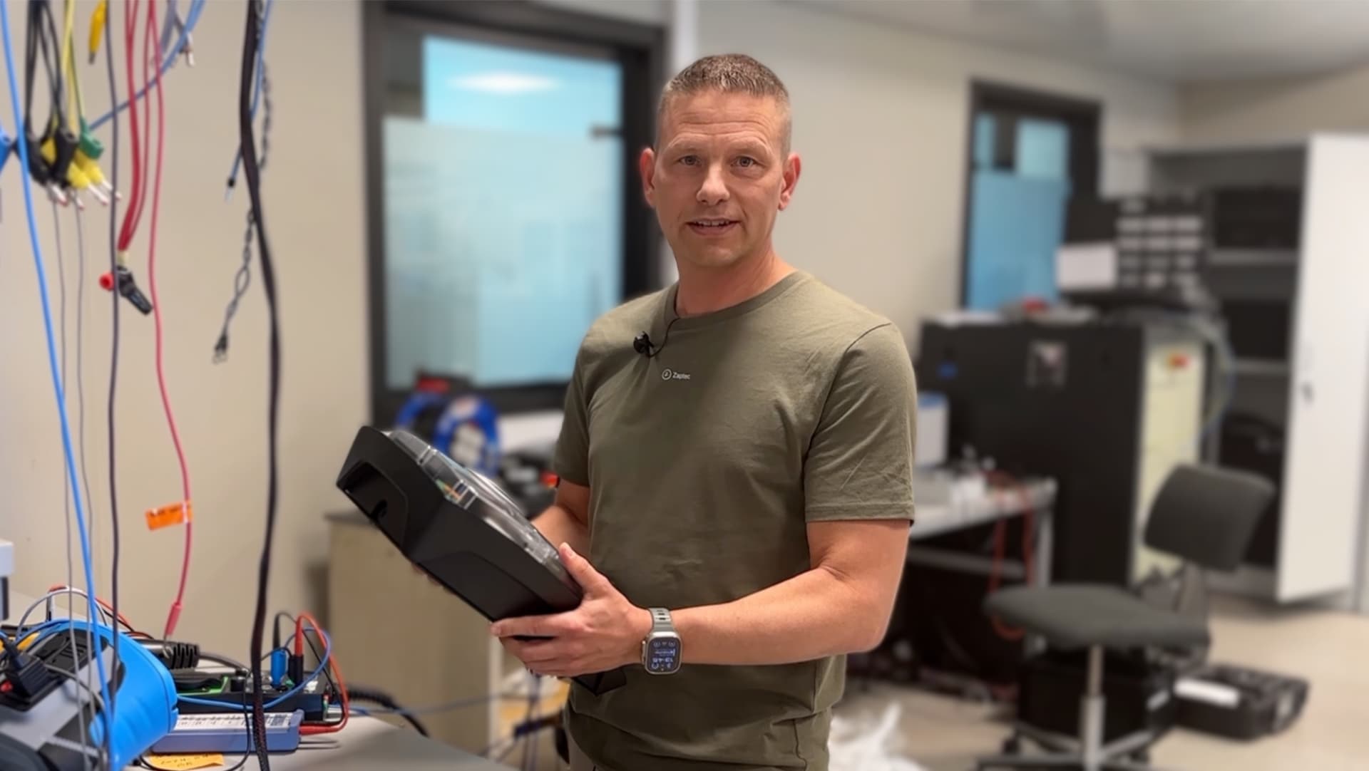 En man i grön t-short och kort mörk frisyr står och håller i en Zaptec Pro laddare i ett testlabb