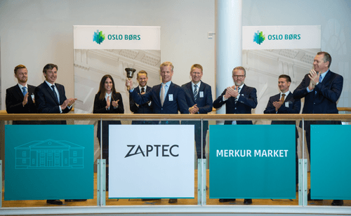 En gruppe representanter fra Zaptec feirer selskapets børsnotering på Oslo Børs. De står i formelle antrekk og applauderer mens den seremonielle bjellen ringes, med skilt som viser «ZAPTEC» og «MERKUR MARKET» i bakgrunnen.
