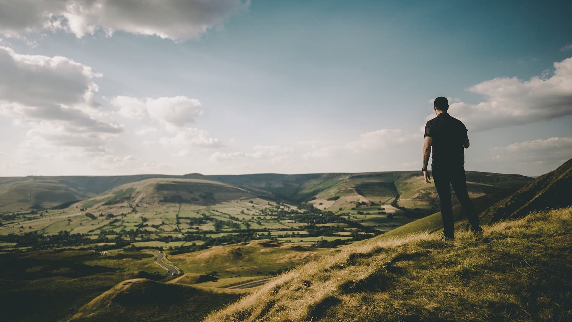 En person i mörka kläder står på toppen av ett berg täckt i gräs och kollar ut över ett grönt landskap