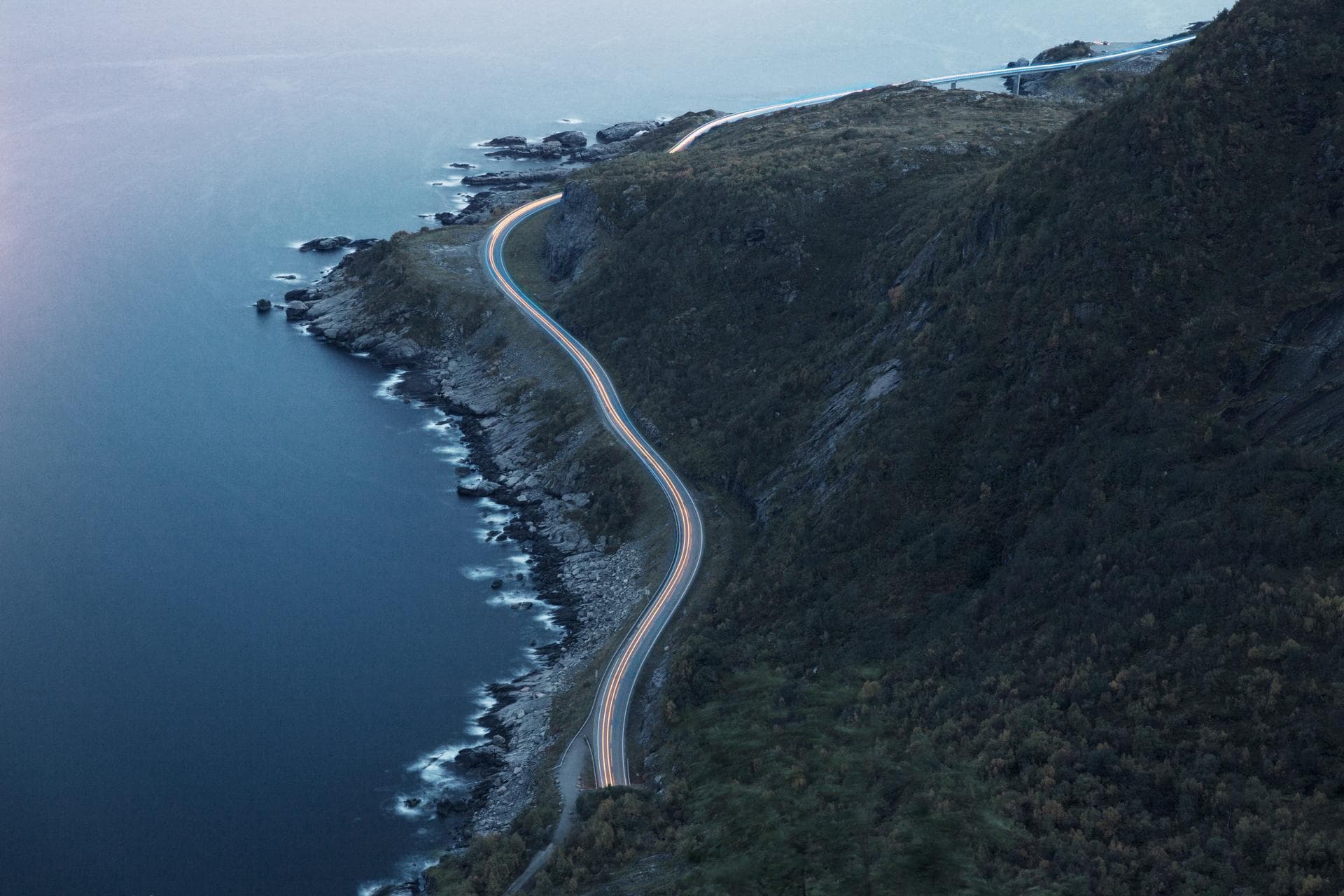 En svingete kystvei følger kanten av en bratt, grønn fjellside, med havet på den ene siden og ulendt terreng på den andre. Scenen er rolig og stemningsfull i mykt kveldslys.
