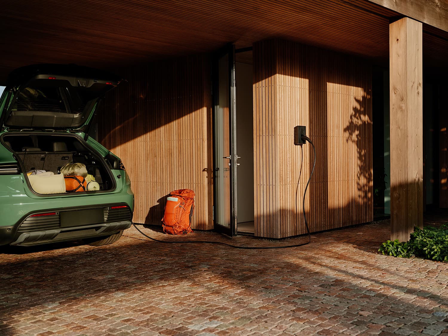 Green car with open trunk charging at Zaptec Go home charger on wooden wall, orange backpack beside, under modern carport.