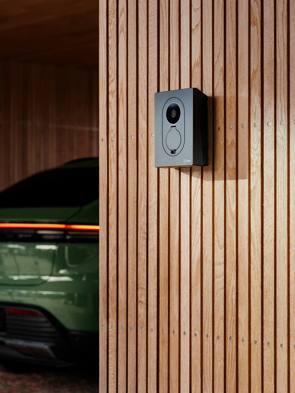 Närbild på Zaptec Go 2 hemmaladdare på trävägg, bakdel av grön bil med rött bakljus synligt i carport.
