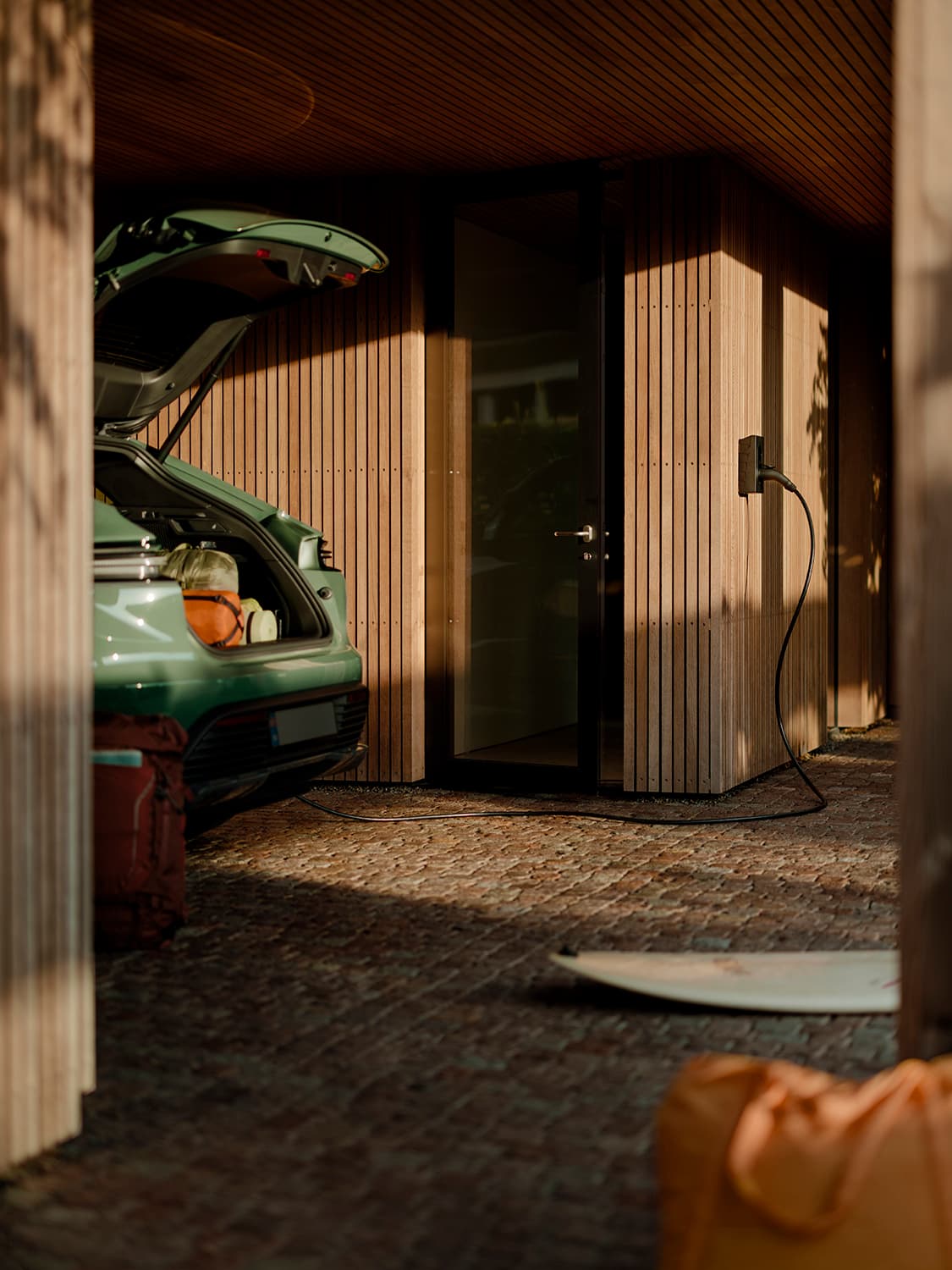 Groene auto met open kofferbak laadt op bij Zaptec Go 2 thuislader aan houten wand, oranje rugzak en surfplank op de voorgrond.