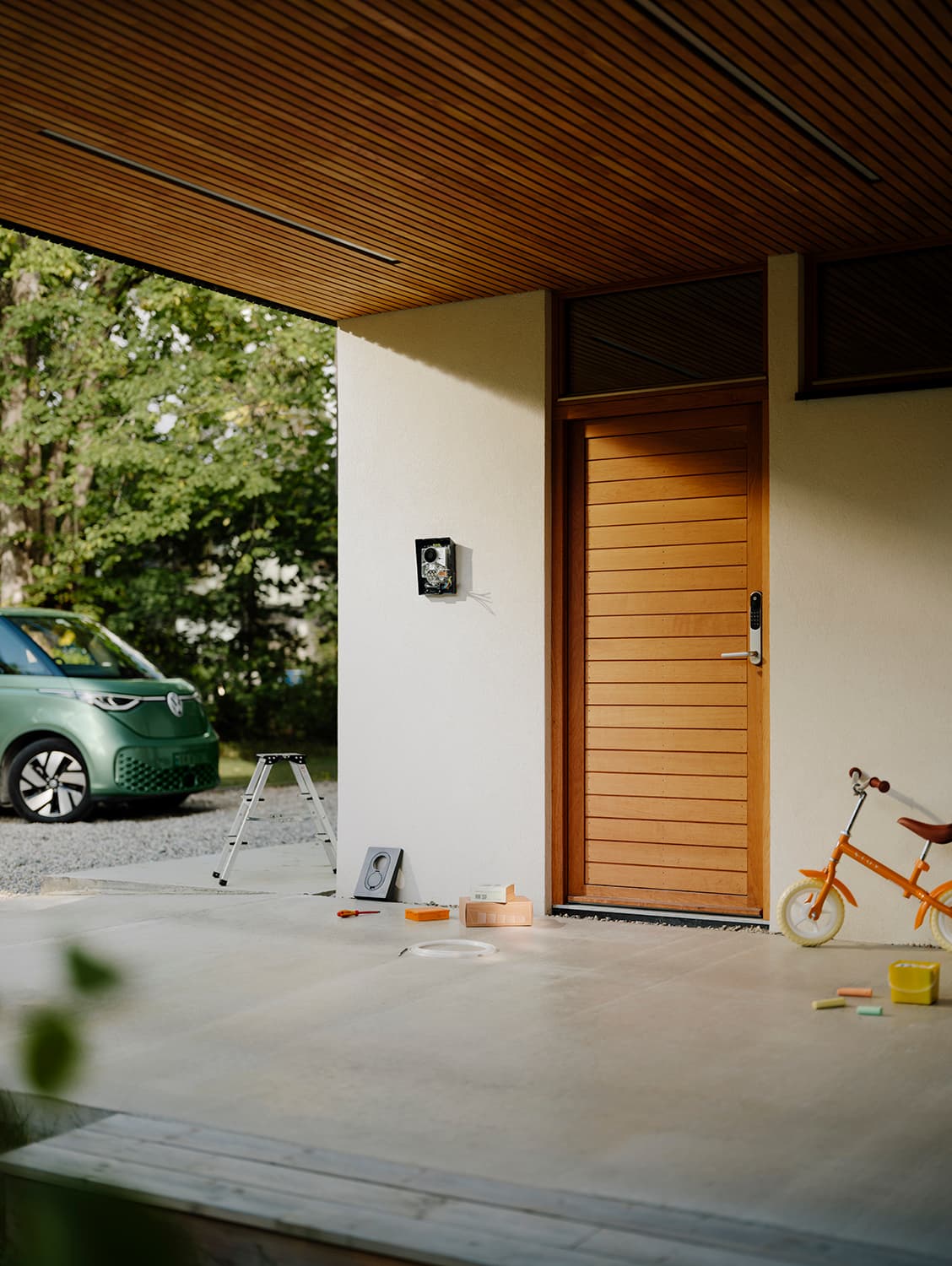 Onvoltooide Zaptec Go 2 thuislader op witte muur bij houten deur, groene bestelwagen, gereedschap en oranje kinderfiets buiten.