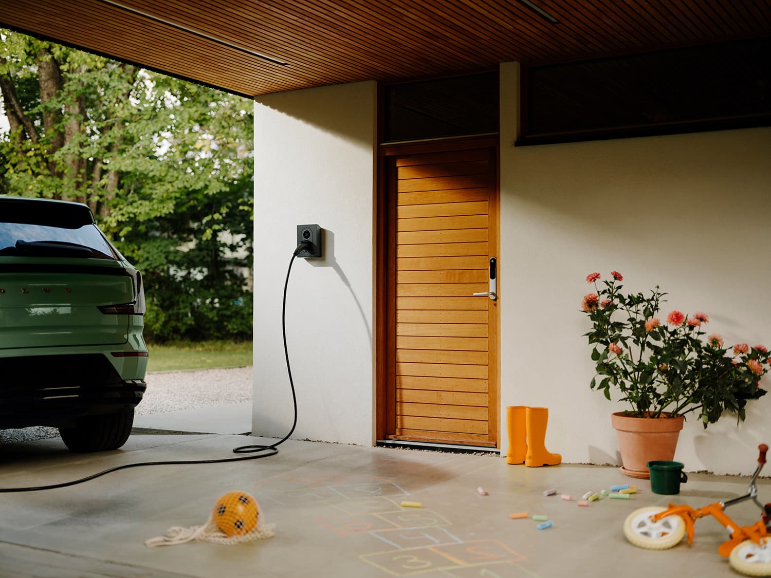 Grønn bil parkert ved Zaptec Go 2 hjemmelader på hvit vegg nær tredør, barneleker og blomster på verandaen.