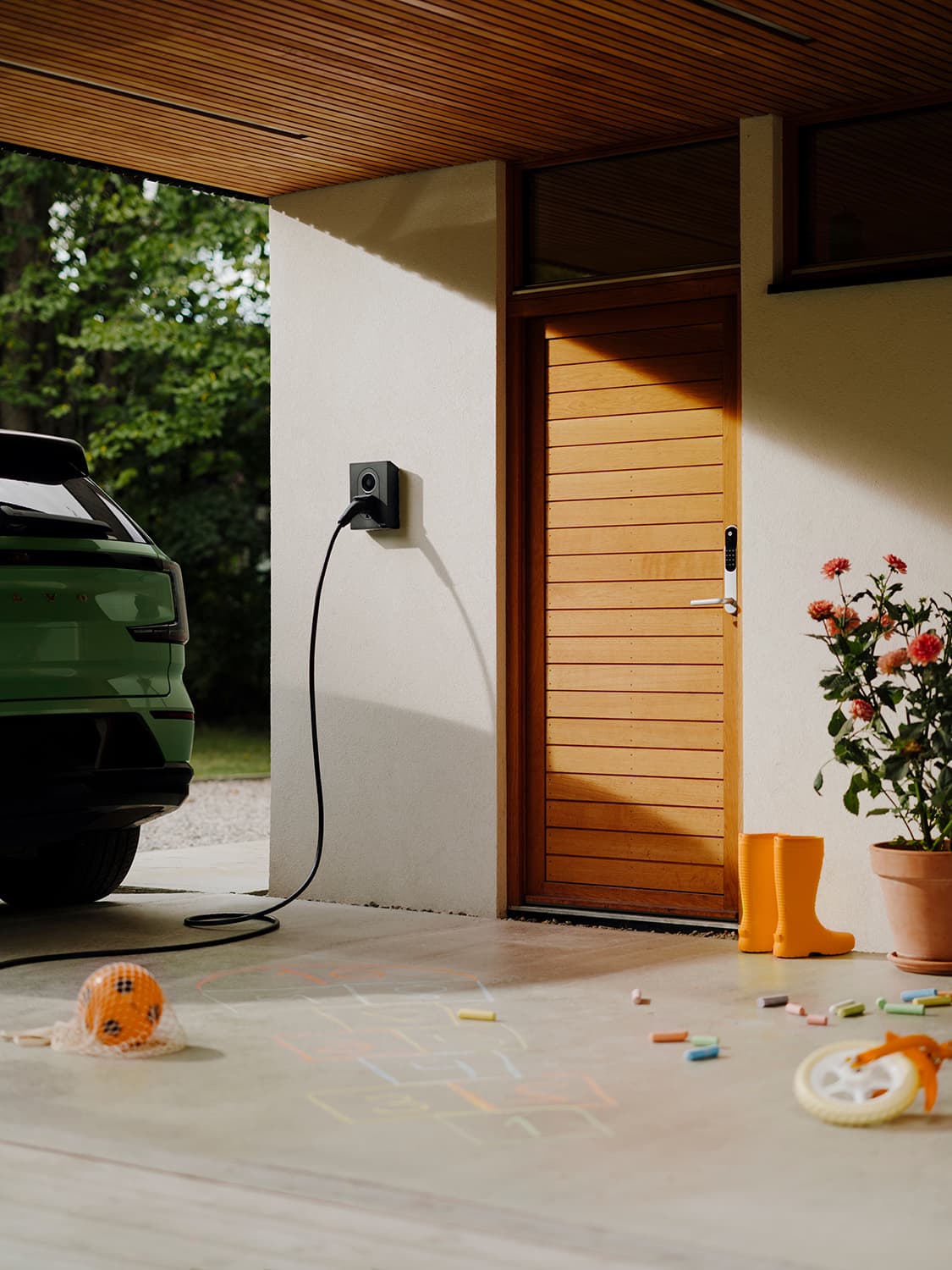 Green car charging at Zaptec Go 2 home charger on white wall, wooden door, children’s chalk drawings and toys on the ground.
