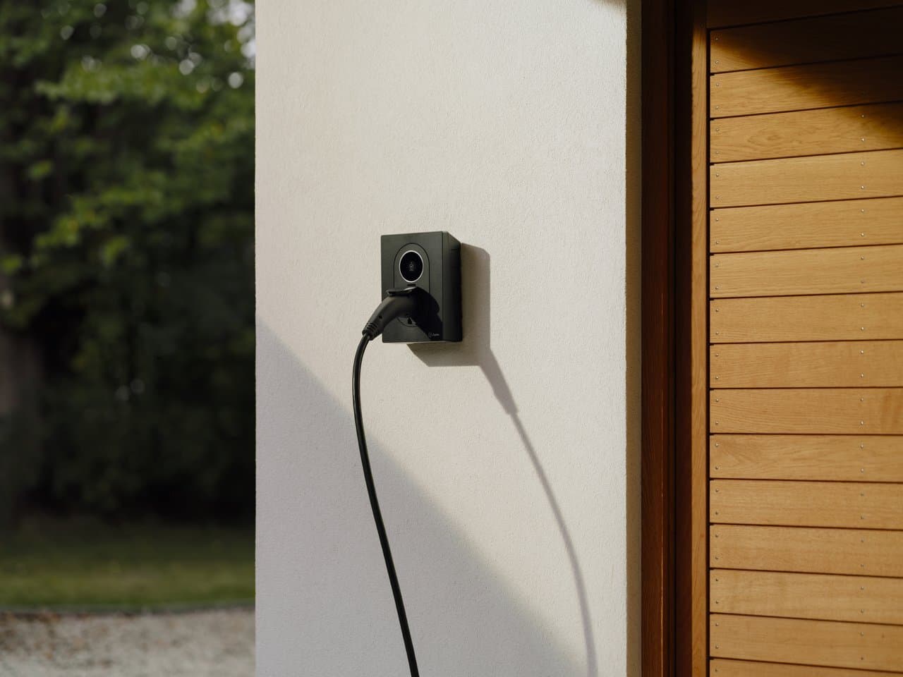 Gros plan sur Zaptec Go 2 borne branchée sur mur blanc près d’une porte bois, soleil et jardin en arrière-plan.